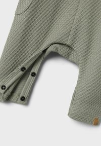 Mono verde con textura de waffle y cierres a presión en las aberturas de las piernas. Presenta una pequeña etiqueta con logo. El material parece suave y transpirable.