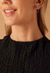 Zilveren hoepeloorbellen met een schakellinkdetail, die een glanzende afwerking tonen. Draag ze met een zwarte blouse met textuur en geplooide stof.