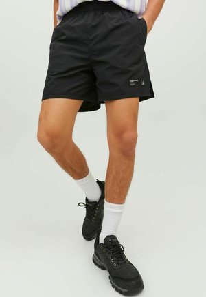 Schwarze Shorts aus leichtem Material mit elastischem Bund und einem seitlichen Logopatch. Kombiniert mit schwarzen Wanderschuhen und weißen Socken.