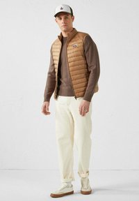 Gilet imbottito beige sopra un maglione marrone, pantaloni bianchi e sneakers color crema. Cappello bianco e marrone con logo. Design semplice, vestibilità casual.