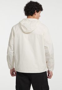 Jachetă cu glugă de culoare deschisă, cu o croială lejeră, realizată dintr-un material texturat, cu manșete elastice și un design simplu. Pantaloni negri prezentați mai jos.