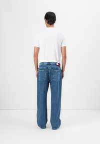 Άν�δρας με λευκό κοντομάνικο πουκάμισο και φαρδιά μπλε τζιν, στέκεται απέναντι σε έναν απλό ανοιχτό γκρι φόντο.