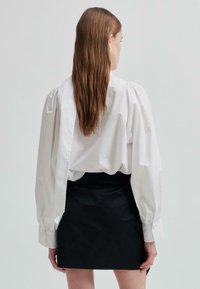 Žena s dlouhými hnědými vlasy, oblečená v bílé blůze s nařasenými rukávy a černé mini sukni, stojící zády k fotoaparátu na jednolitém pozadí.