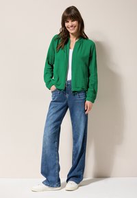 Groene textuur bomberjack met geribde boorden, gecombineerd met loszittende blauwe jeans en witte sneakers, tegen een neutrale achtergrond.