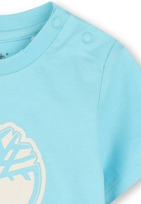 Lichtblauw t-shirt met een ronde hals, korte mouwen en twee drukknoopjes op de schouder. Heeft een wit grafisch ontwerp.