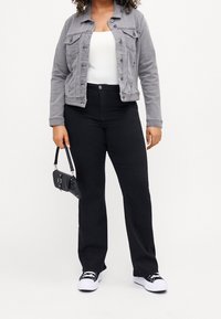 Žena v šedé džínové bundě, bílé blůze, černých vysokých džínách, černo-bílých teniskách, držící malou černou kabelku po boku.