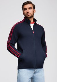 Marineblaue Jacke mit Reißverschluss und roten Seitenstreifen sowie Schriftzug, aus dehnbarem Material gefertigt; verfügt über einen hohen Kragen und zwei Vordertaschen, schlanke Passform.