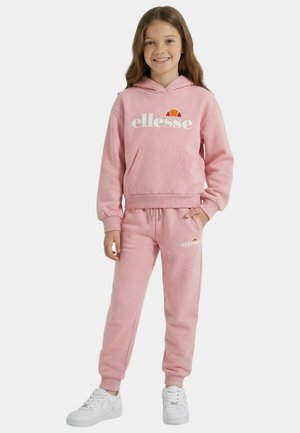 Jeune fille souriante, portant un sweat à capuche ellesse rose et un pantalon de jogging assorti avec des baskets blanches, debout devant un fond uni.