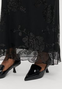 Melna grīdas garuma kleita ar ziedu aplikācijām no fliteriem un caurspīdīgu audumu. Melni lakotas ādas apavi ar smailu purngalu un maziem papēžiem.