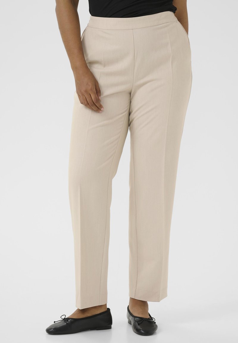 Beige tailored pants with a smooth texture, straight legs, and a mid-rise waistband. Paired with black flats featuring bow accents.