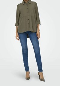 Camicia verde oliva con bottoni, colletto, maniche arrotolate e un orlo posteriore più lungo, abbinata a jeans skinny blu e tacchi alti taupe.