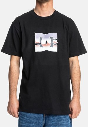 Mand iført sort t-shirt med stiliseret hvidt og pink DC-logo, kombineret med blå jeans, stående mod en ensfarvet baggrund.