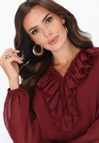 Bordó blúz fodros nyakkivágással és fodros ujjakkal, könnyű anyagból készült. A modell az arcához emeli a kezét, arany karika fülbevalót visel.
