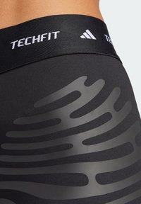 Detailní záběr na černé sportovní legíny s TECHFIT pasem a reflexním abstraktním vlnitým vzorem na boku.