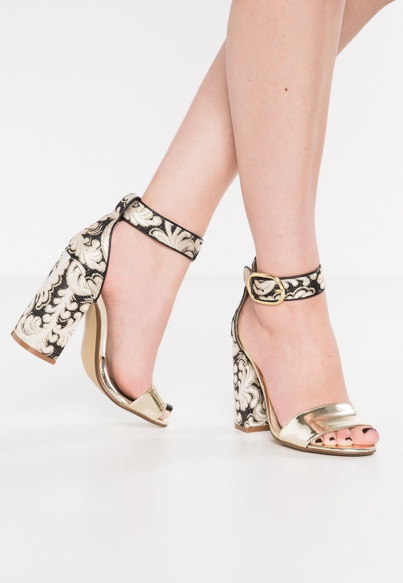 Pieds portant des talons hauts ouverts dorés avec des brides de cheville brodées noires et dorées et des talons blocs sur un sol blanc.