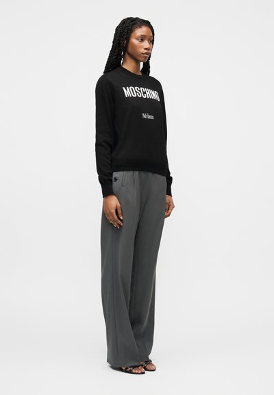 Femme aux longs cheveux tressés portant un sweat-shirt noir Moschino Milano, un pantalon large gris et des sandales noires à bout ouvert.