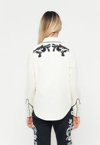 White long-sleeve shirt with black embroidered floral design on back, rounded hem, contrasting collar, and button accents on cuffs.
