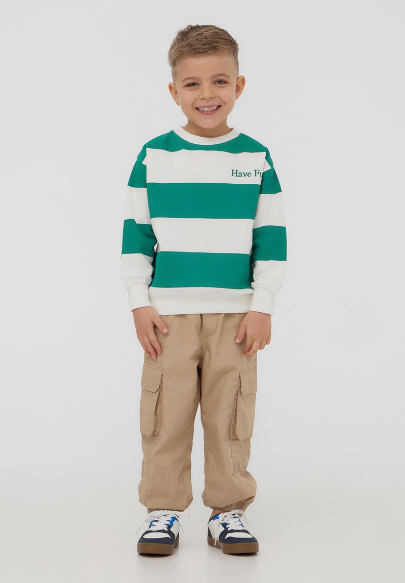 Felpa a strisce verdi e bianche con testo "Divertiti", abbinata a pantaloni cargo beige e sneakers bianchi con accenti blu e neri.