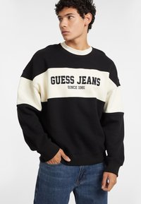 Schwarzer und cremefarbener Sweatshirt mit dem Text "GUESS JEANS" in Fettdruck. Besitzt einen Rundhalsausschnitt, lange Ärmel und einfarblich abgestimmtes Design.