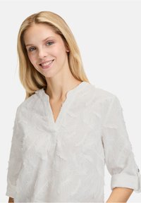 Blusa bianca con colletto alla mandarina, caratterizzata da motivi a foglia testurizzati e maniche a tre quarti, realizzata in tessuto leggero.
