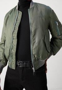 Blouson aviateur vert avec fermeture éclair à l'avant, col gris, poignets en côtes et détails de poches latérales ; porté par-dessus un col roulé noir et un jean noir.