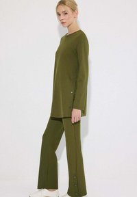 Γυναίκα που φορά ζωντανή πράσινη μακριά τουνίκ με μακριά μανίκια και ταιριαστά παντελόνια με καμπάνα και λεπτομέρειες με κουμπιά, στέκεται μπροστά σε απλό λευκό φόντο.