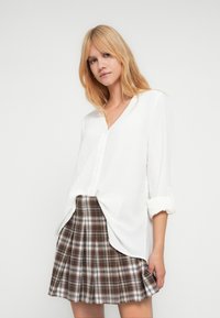 Chemise blanche boutonnée avec une finition texturée et des manches retroussées, associée à une jupe plissée à carreaux marron avec des lignes blanches, marron et noires.