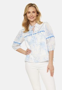 Blusa bianca con stampa floreale blu, dotata di colletto, maniche a tre quarti e un dettaglio con fiocco sullo scollo. Abbinata a pantaloni bianchi.
