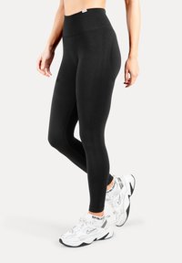 Schwarze, hoch taillierte Leggings aus elastischem Stoff mit glatter Textur und figurbetontem Design, kombiniert mit weißen Sportschuhen.