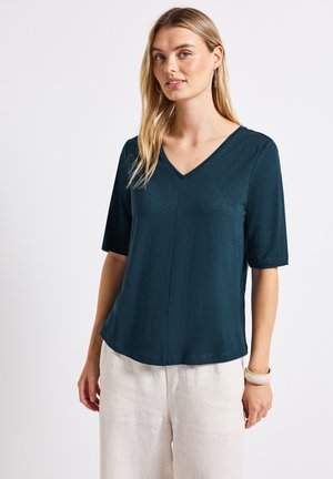 Blouse turquoise à col en V avec des manches courtes, texture lisse et coupe décontractée. Présente un détail de couture sur le devant et se marie bien avec un pantalon clair.