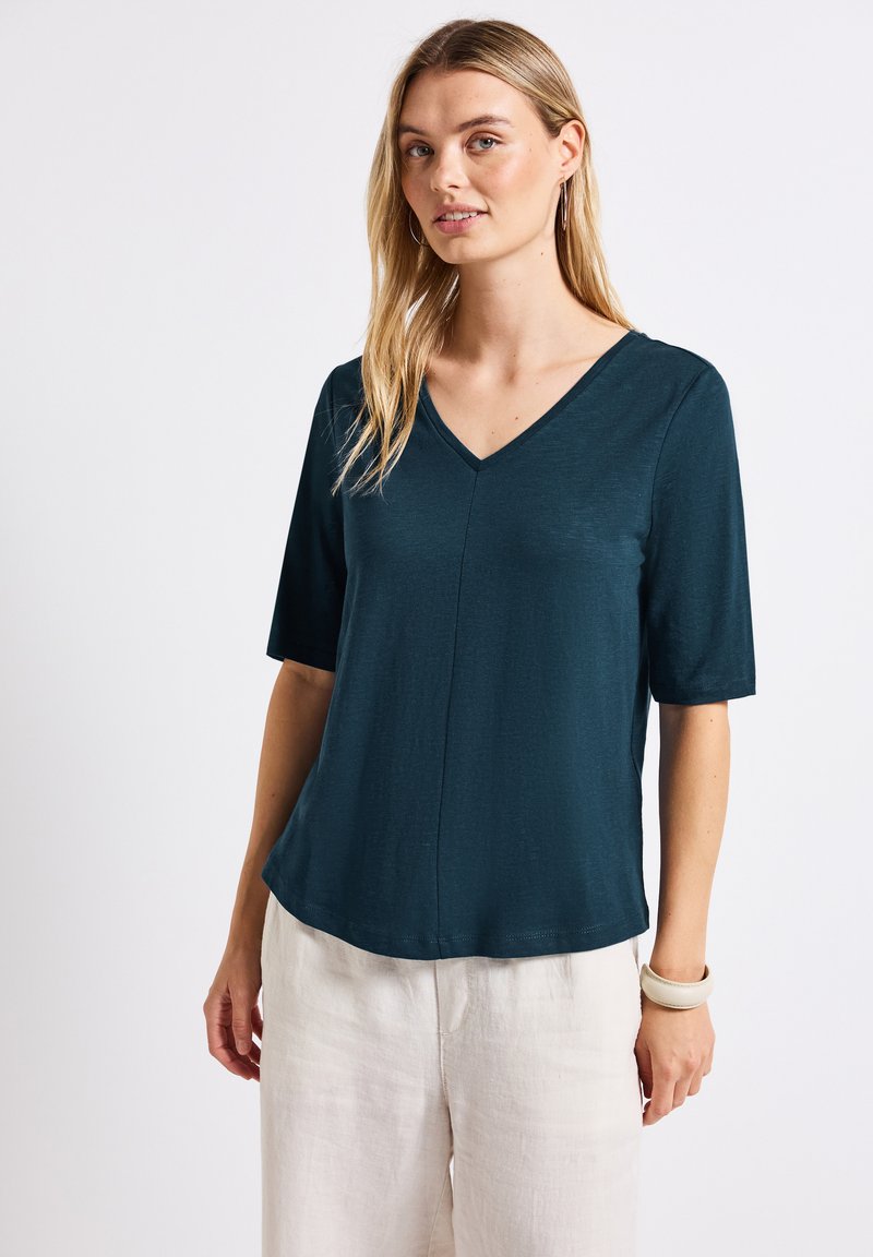 Tyrkysová blůza s výstřihem do V a krátkým rukávem, hladká textura a uvolněný střih. Má ševový detail na přední straně a skvěle se hodí k světlým kalhotám.