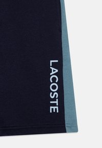 Jūras zila auduma šorti ar gaiši zilām sānu joslām, uz kuriem vertikāli pa kreiso pusi ir balts "LACOSTE" logotipa teksts.