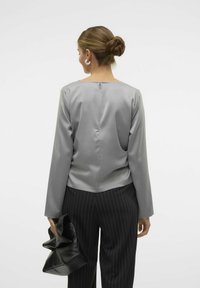 Top lungo a maniche con zip sul retro in satin grigio, abbinato a pantaloni a righe, mentre tiene una borsa texture nera.