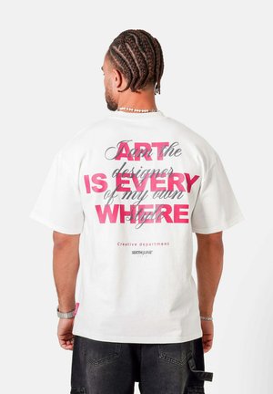 Hombre con trenzas que lleva una camiseta blanca con texto rosa y negro, combinado con pantalones cortos de mezclilla negros, visto desde atrás.