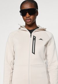 Lichtbeige sweater met rits en capuchon, met zwarte accenten en een voorzak met rits. Draag het met donkere oversized zonnebrillen.