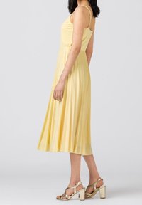 Gul plisserad midiklänning med tunna axelband och figurnära överdel, kombinerad med metalliska guldklackar. Slät textur, flödande tyg.