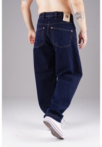 Dunkelblaue Jeans aus Denim mit lässiger Passform, ausgestattet mit fünf Taschen, kupfernen Nieten und einem tanfarbenen Lederlogo-Patch auf der Rückseite.