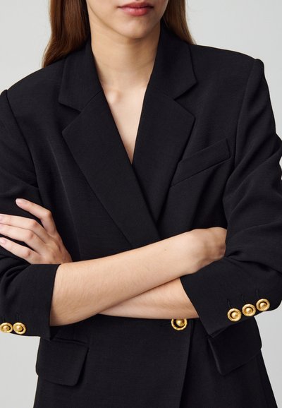 Blazer negro entallado con diseño de escote en V, que presenta detalles de botones dorados en los puños y un bolsillo en el pecho. Tejido texturizado.