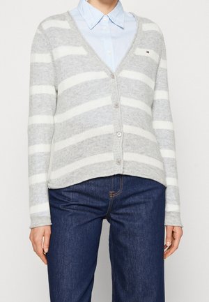 Vrouw met grijs-wit gestreepte cardigan over een lichtblauw overhemd, met donkerblauwe spijkerbroek, staand tegen een effen achtergrond.