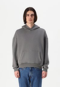 Hanorac gri din material amestec de bumbac, cu buzunar frontal, glugă cu șnur și manșete strâmte, asortat cu blugi denim albastre.