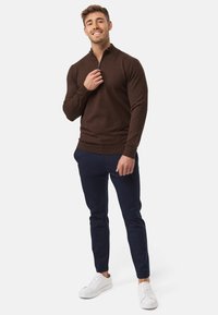 Suéter marrón con cierre, de manga larga, con un cuerpo ajustado, combinado con pantalones tapered azul marino y zapatillas blancas. Textura suave, diseño casual.