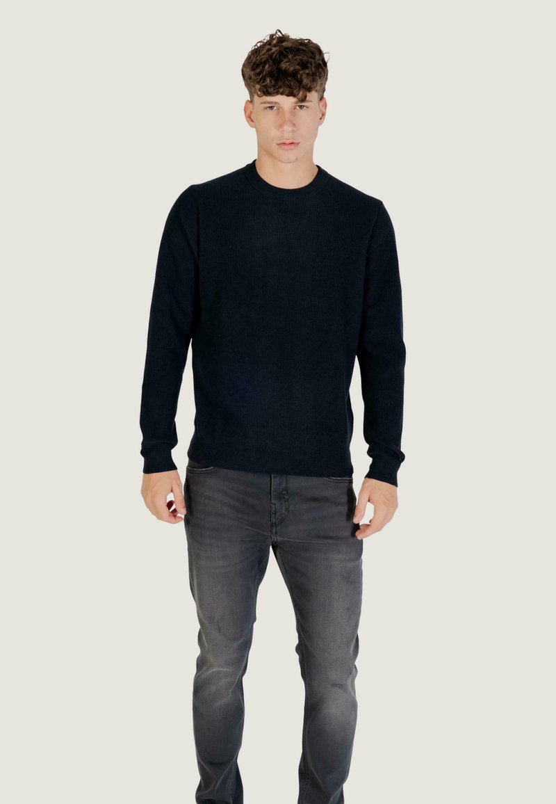 Tamna mornarska pamučna majica s rebrastim uzorkom i okruglim izrezom, u kombinaciji s sivim trapericama. Tkanina izgleda mekano, s dugim rukavima i uskim krojem.