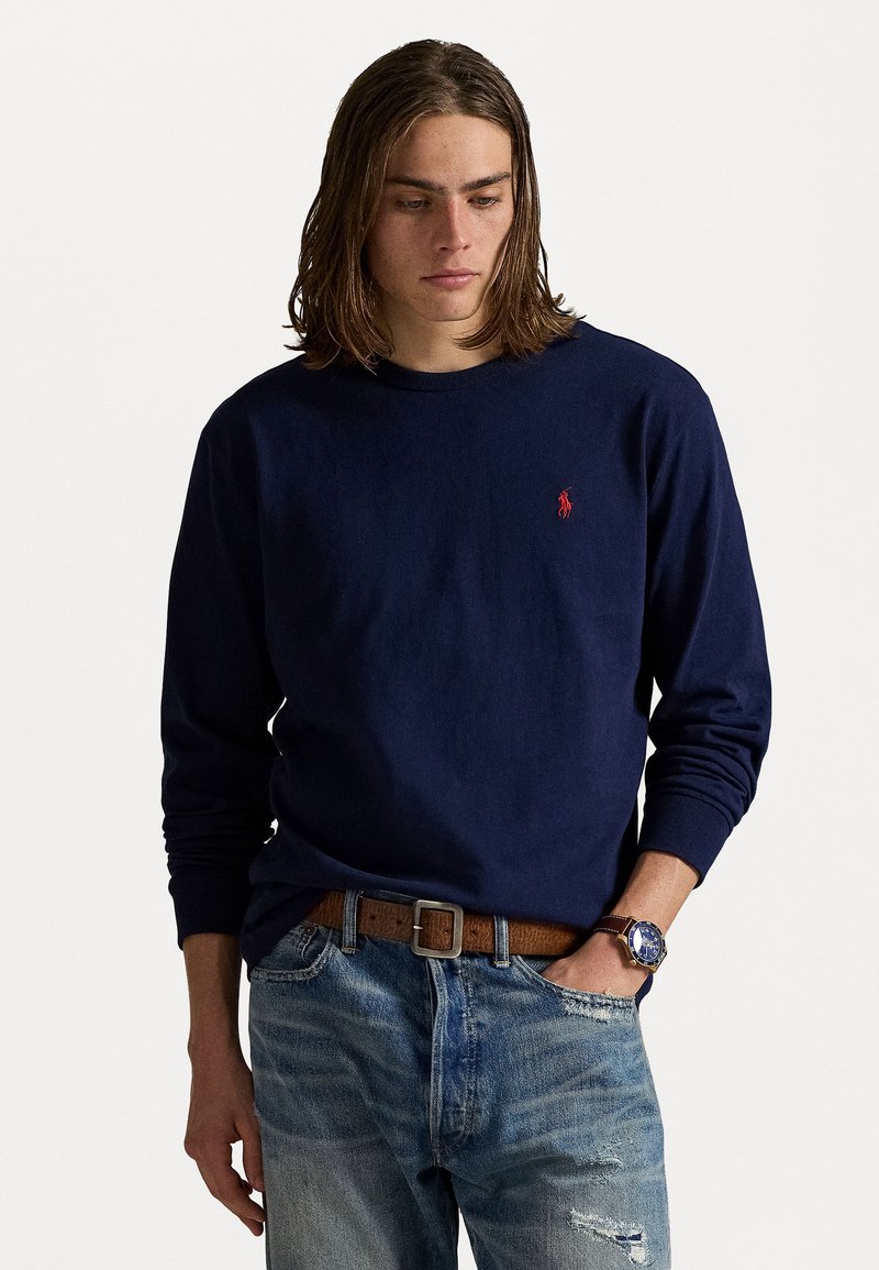 Marineblauwe lange mouwen t-shirt, gemaakt van katoen, met een ronde halslijn, heeft een klein rood logo op de linkerborst; gedragen met lichtgewassen versleten jeans en een bruine riem.