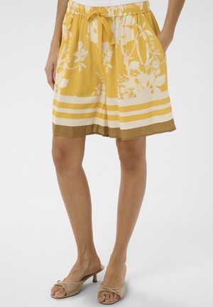 Shorts jaunes à fleurs arrivant aux genoux avec une taille à cordon et des rayures horizontales blanches et marron, portés avec des sandales à talons beige.