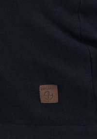 Granatowy materiał o gładkiej teksturze z kwadratową brązową skórzaną metką wytłoczoną z napisem "KEY LARGO" oraz okrągłym wzorem logo.