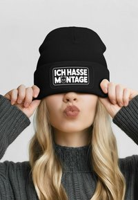 Black knit beanie with a rectangular patch displaying "ICH HASSE MONTAGE" and a cat design. Worn low over the forehead.
