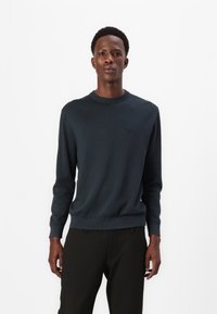 Dunkelblauer Pullover mit runder Ausschnitt, gerippten Bündchen und Saum. Weicher Stoff, minimales Logodesign auf der Brust, kombiniert mit gestreiften schwarzen Hosen.