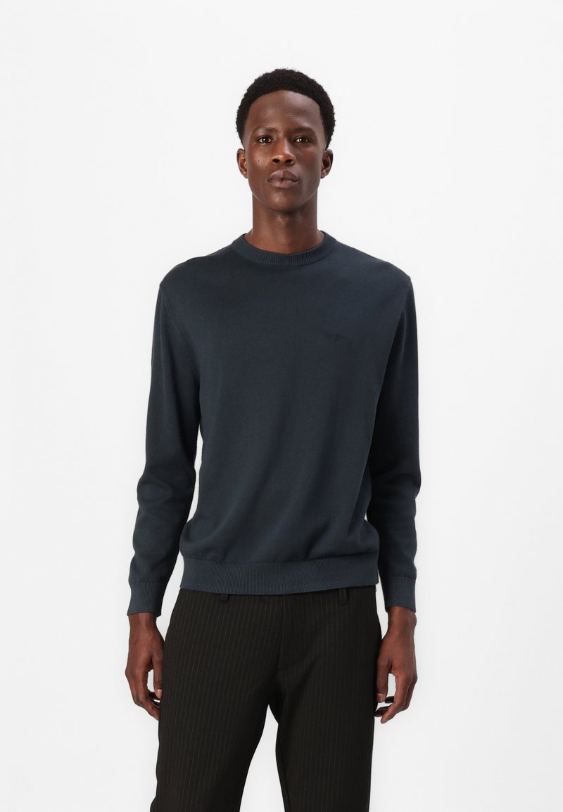 Dunkelblauer Pullover mit runder Ausschnitt, gerippten Bündchen und Saum. Weicher Stoff, minimales Logodesign auf der Brust, kombiniert mit gestreiften schwarzen Hosen.