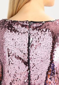 Vista posteriore di una persona che indossa un top fucsia con paillettes e una cerniera nera che scende lungo il centro della parte superiore della schiena.