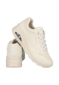 Zapatillas deportivas de color beige con una parte superior sintética lisa, detalles perforados, sistema de cordones y una suela acolchada que cuenta con una unidad de aire visible.